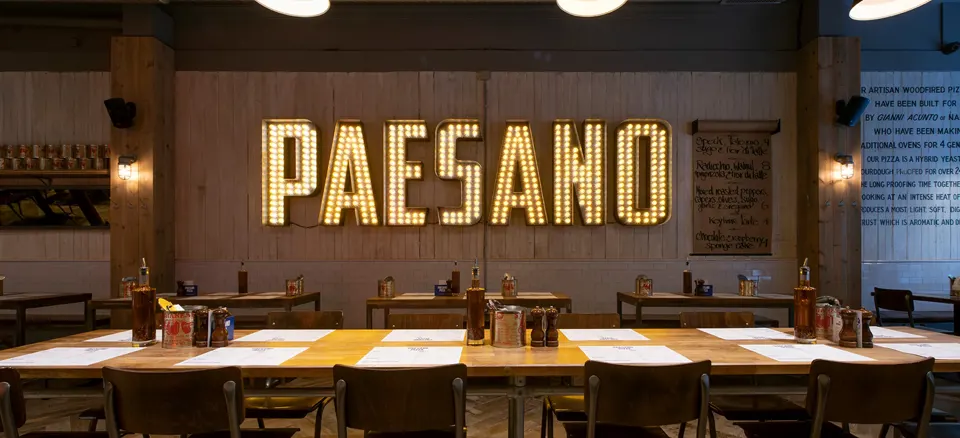 Inside a Paesano Pizza restaurant, with the illuminated Paesano sign above long tables