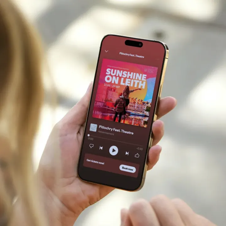 A hand holding a phone showing a Pitlochry Festival Theatre 'Sunshine on Leith' audio advert