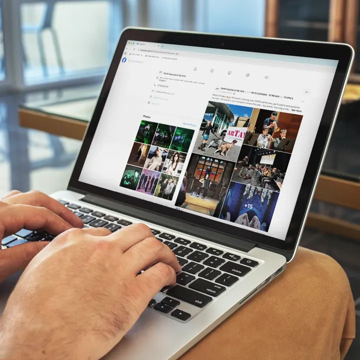 A person's hands typing on a laptop displaying the Facebook page of the Perth Festival of the Arts, featuring various event photos.