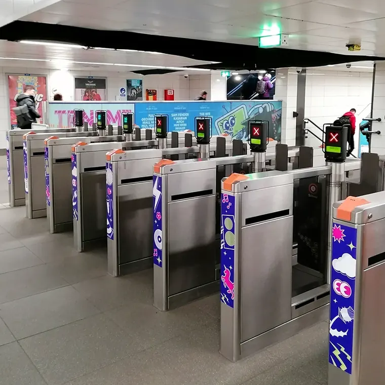 A row of stainless steel subway ticket barriers decorated with colourful TRNSMT decals. In the background, the large TRNSMT festival advertisement is visible behind the gates.
