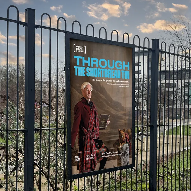 A printed poster in a black frame, on a fence, advertising the National Theatre of Scotland's show Through The Shortbread Tin