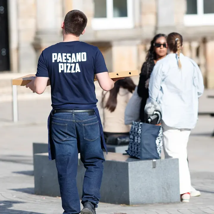 A Paesano Pizza staff member carrying pizza boxes across a city square