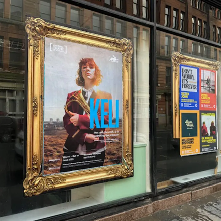 A printed poster for the National Theatre of Scotland production of Keli displayed inside a gold frame on a storefront window in Glasgow.