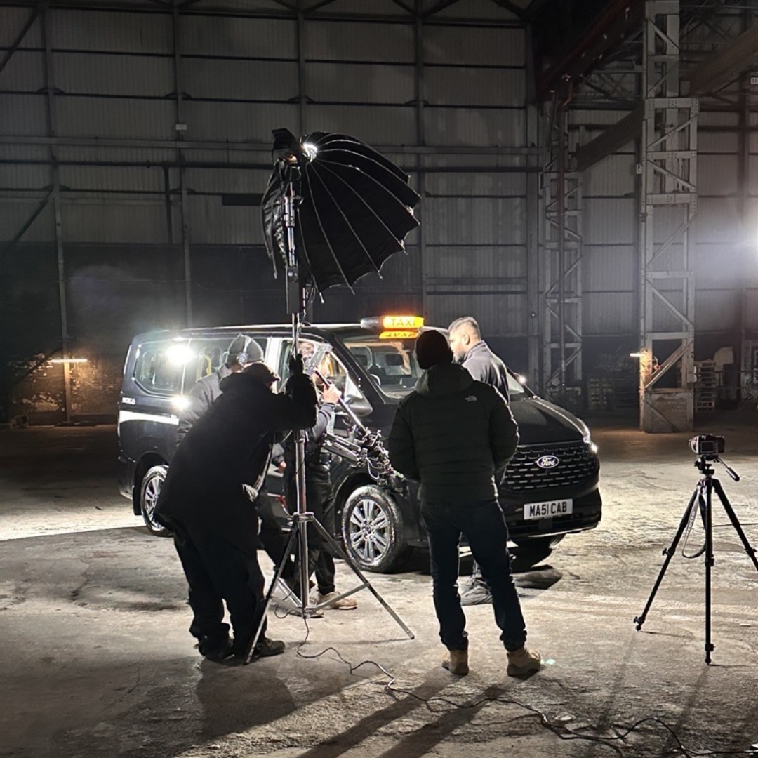 Film crew setting up lights and cameras around a MAXICab taxi in a warehouse