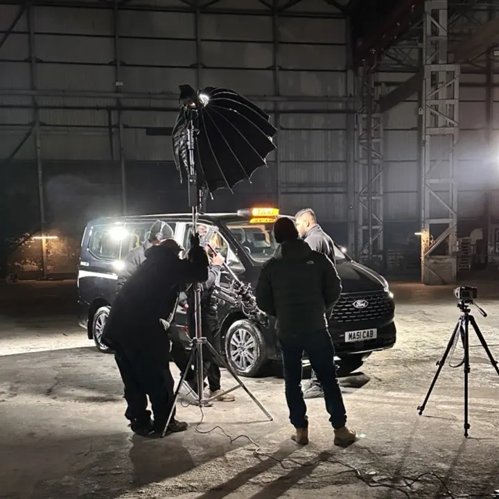 Film crew setting up lights and cameras around a MAXICab taxi in a warehouse