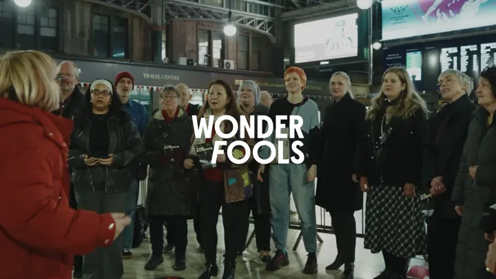 Wonder Fools logo over a community choir singing in Glasgow Central Station