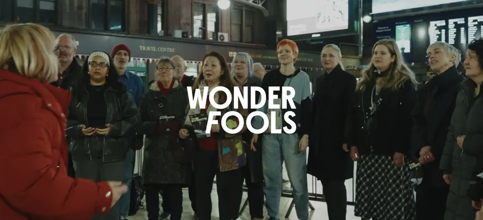 Wonder Fools logo over a community choir singing in Glasgow Central Station