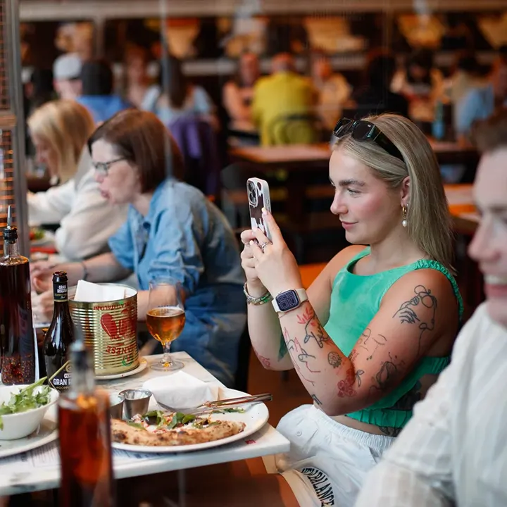 A woman at Paesano Pizza takes a photo of her dining companion. On the marble table sits a Neapolitan pizza and a cold glass of beer.