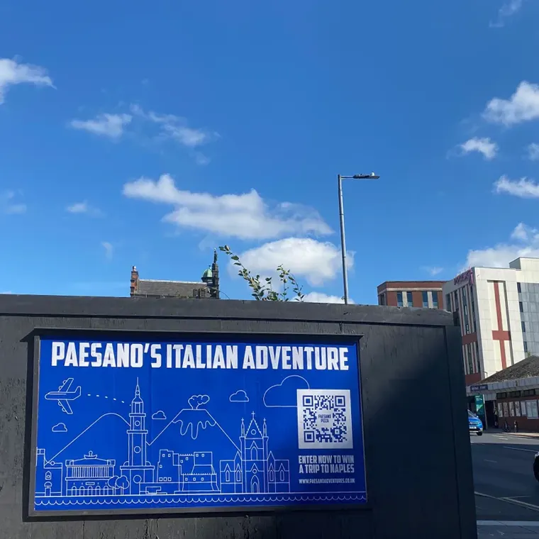 A 'Paesano's Italian Adventure' billboard on a Glasgow street under a blue sky