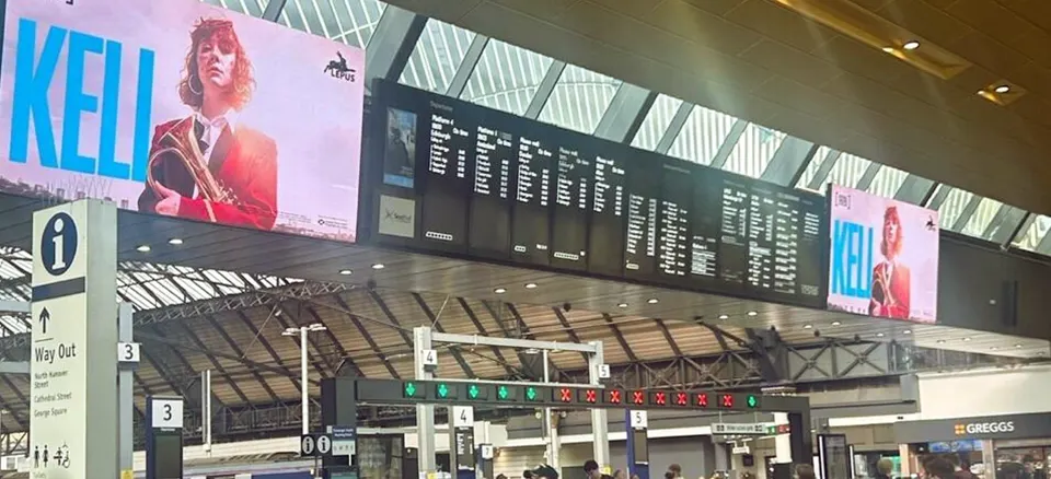 A digital billboard inside Glasgow Queen Street train station displays a poster for the National Theatre of Scotland's production of Keli for dates June 11–14 at Tramway, Glasgow.