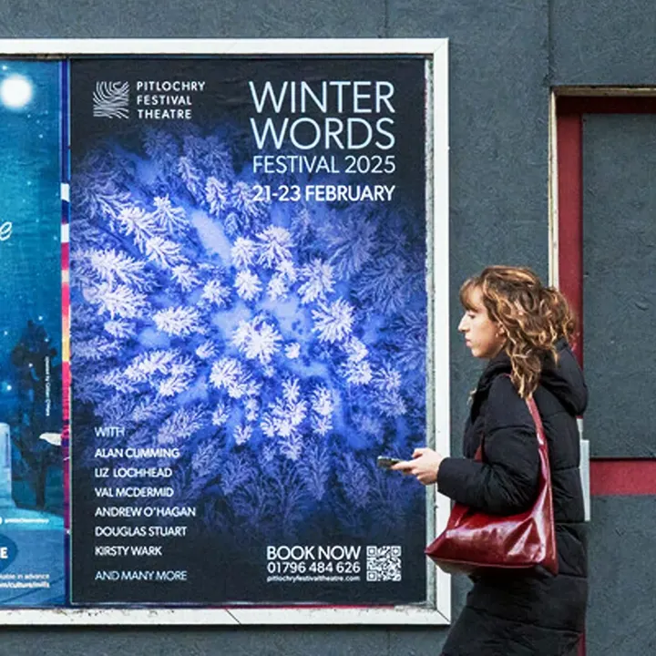 A woman walking past a 'Winter Words Festival 2025' poster listing Alan Cumming, Liz Lochhead and Val McDermid
