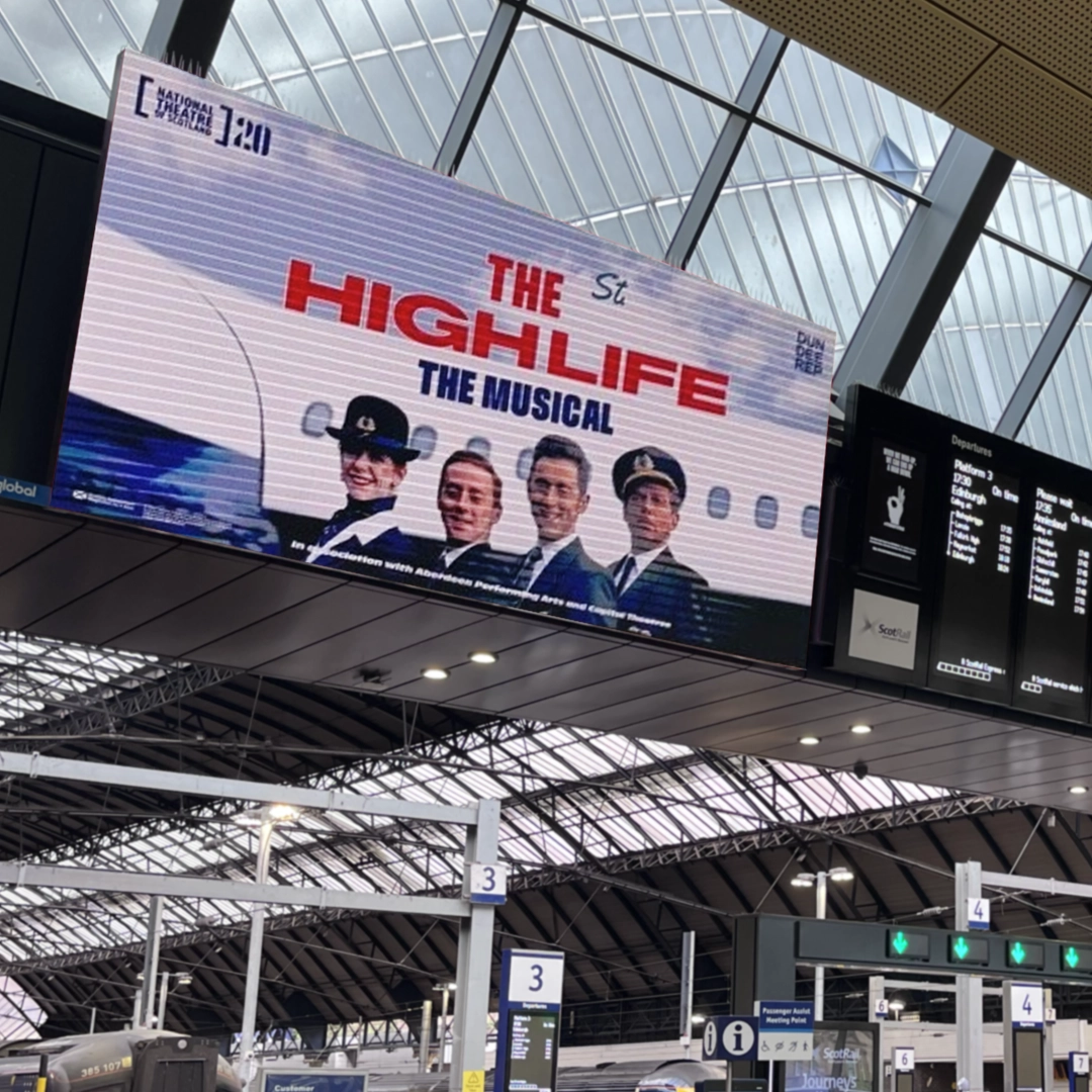 A large screen at Glasgow Central Station showing 'The High Life: The Musical' advert