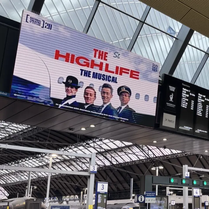 A large screen at Glasgow Central Station showing 'The High Life: The Musical' advert