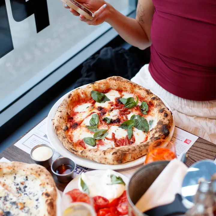 A person at Paesano Pizza holds a phone over a table with a fresh Neapolitan pizza topped with basil, as well as a side of tomatoes and mozzarella, small dipping sauces, and a glass of orange spritz.