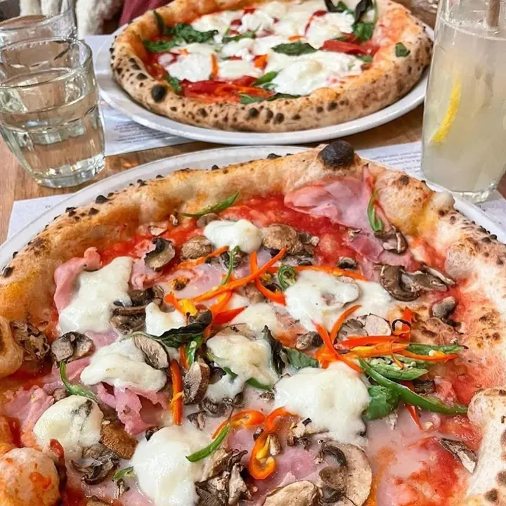 Two Paesano pizzas on a table, one topped with ham, mushrooms and chilli