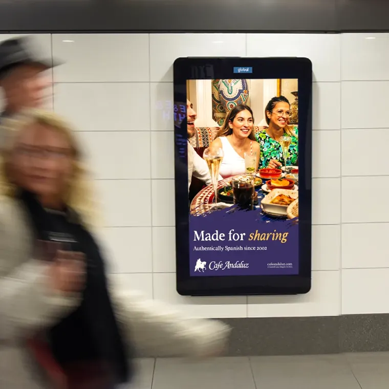 A digital poster in a station passageway showing friends dining at Cafe Andaluz: 'Made for sharing'