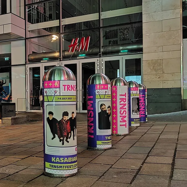 A series of silver-topped bollards on a paved city street in front of an H&M store. The bollards are wrapped in vibrant purple and pink TRNSMT festival advertisements featuring photos of artists like Kasabian and The 1975.