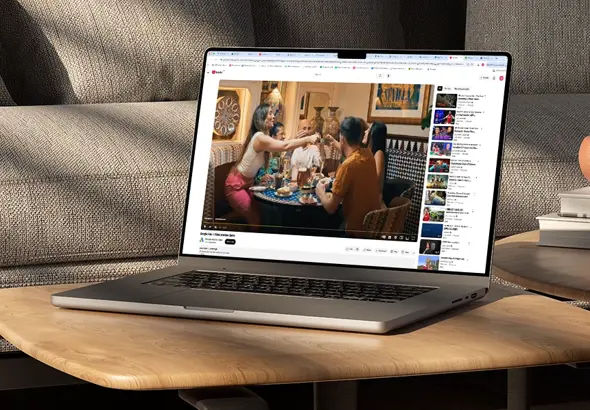A silver laptop sitting on a wooden coffee table in a living room, playing a YouTube video of people dining together.
