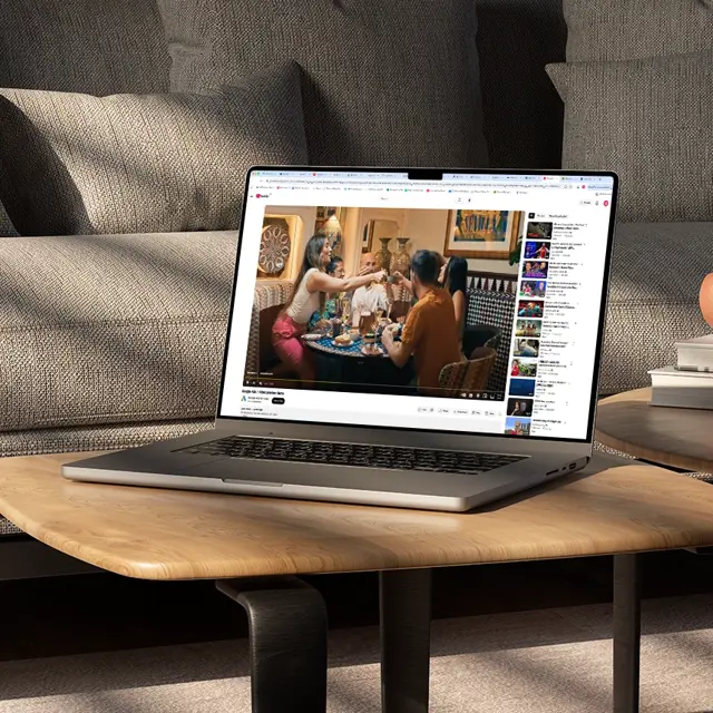 A silver laptop sitting on a wooden coffee table in a living room, playing a YouTube video of people dining together.