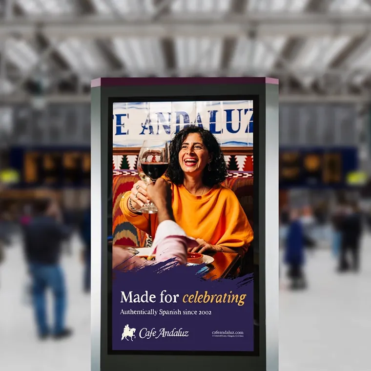 A digital screen at Glasgow Central Station showing a laughing woman with a glass of wine: 'Made for celebrating', Cafe Andaluz