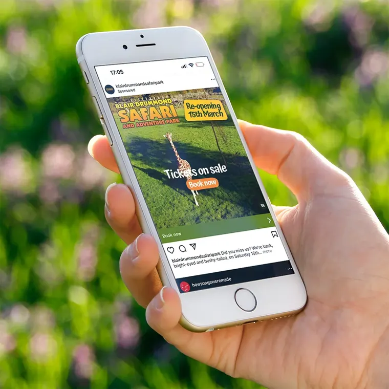 A hand holding a mobile phone showing a Blair Drummond Safari Park sponsored ad, promoting their tickets on sale as the park reopens on the 15th March